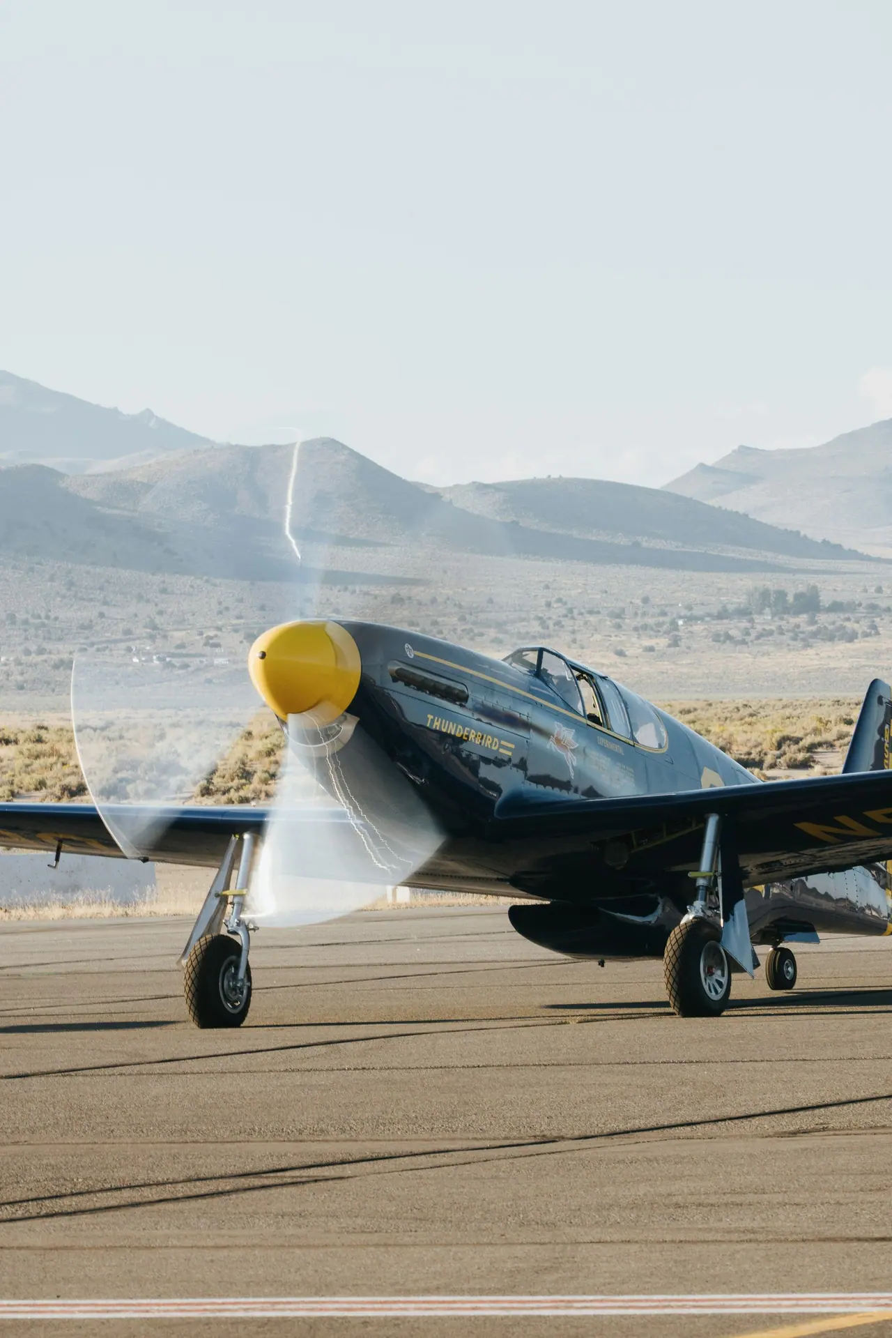 Single-engine piston aircraft with a yellow spinner, propeller turning, on a ramp with hills behind